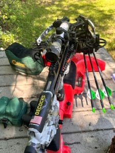 Camouflage compound bow with scope and sight, arrows in a quiver, and a green Gear bag nearby on a wooden surface.