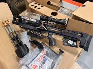 Ravin R470 crossbow with scope and orange-fletched arrows in a cardboard box, surrounded by packing material and accessories.