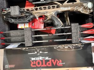 Camouflage-patterned crossbow in foam packaging with a scope and orange bolts nearby.