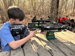 Young boy in a blue shirt aiming a camouflaged crossbow with a scope on a wooden outdoor platform, forest in the background.