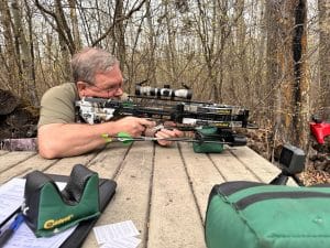 Man lying prone in a forest, aiming a camouflaged crossbow with a scope, surrounded by archery gear and bags.