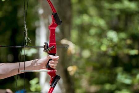 Person holds and draws a red compound bow outdoors, arrow not nocked, in a forest setting.