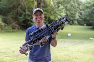 Smiling woman outdoors holding a large camouflage archery bow across her chest in a grassy park setting.