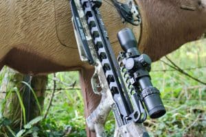 Close-up of a camouflage compound bow with sight and scope mounted on a tree trunk outdoors.