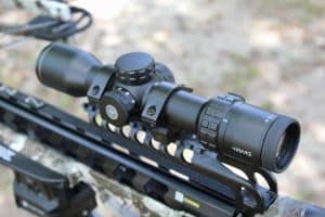 Close-up of a black riflescope mounted on a tactical rifle, outdoors on rocky terrain, with adjustment turrets visible.