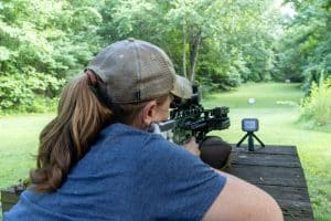 Person with ponytail wearing a mesh cap aims a scoped rifle from a wooden shooting bench in a grassy, tree-lined outdoor range.