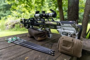 Crossbow with scope on a wooden table outdoors, with arrows laid out nearby in a forest setting