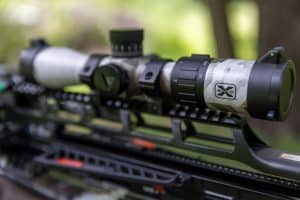Close-up of a rifle scope mounted on a tactical rifle, showing adjustment turrets and a camouflage-patterned magnifier, with a blurred outdoor background.