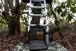 Ladder leaned against a tree in a forest; a backpack is open on a camouflage mat with gear and a thermos nearby.