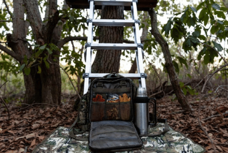 Ladder leaned against a tree in a forest; a backpack is open on a camouflage mat with gear and a thermos nearby.