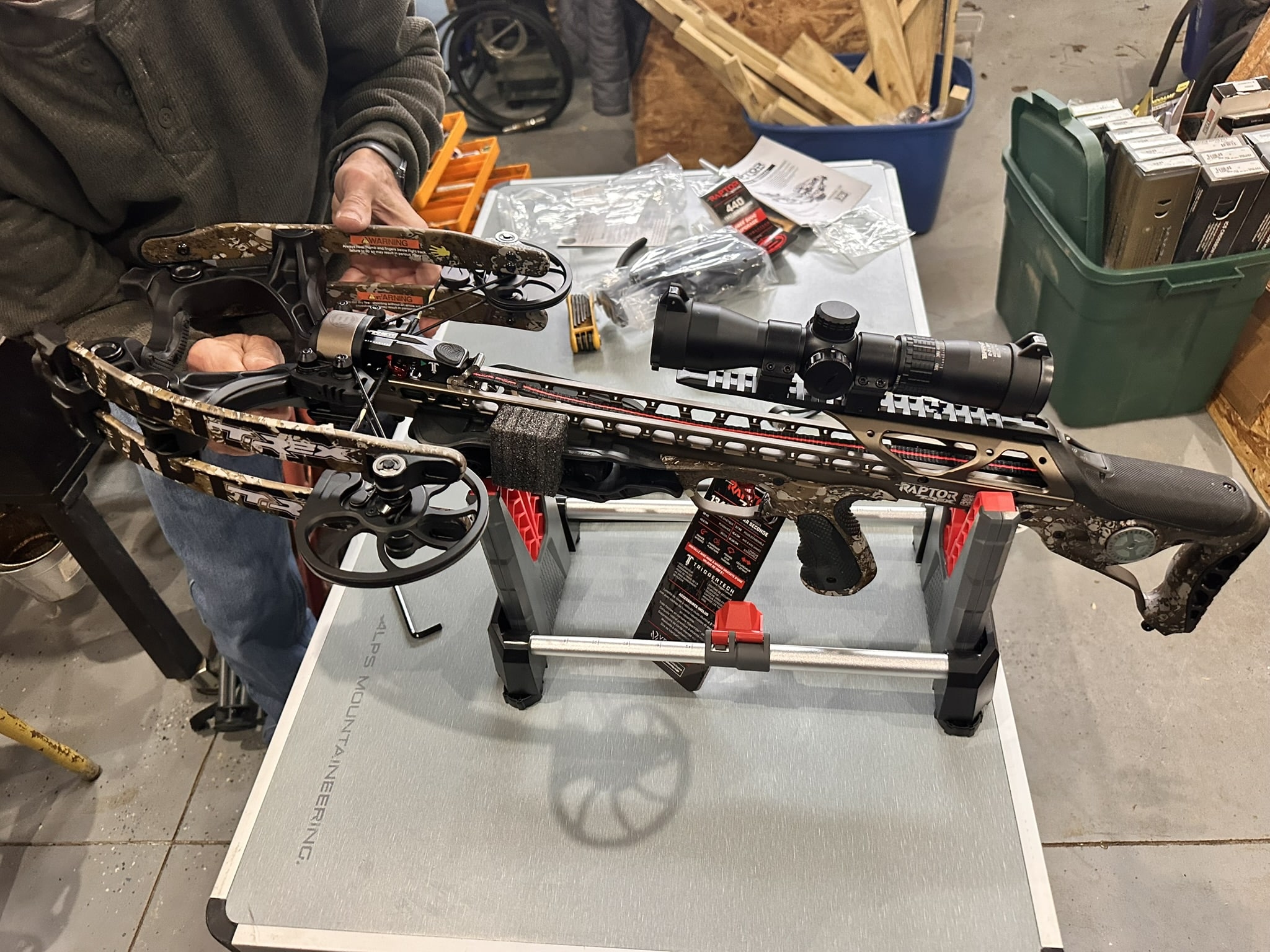 Person inspecting a camouflage crossbow mounted on a work table in a workshop.