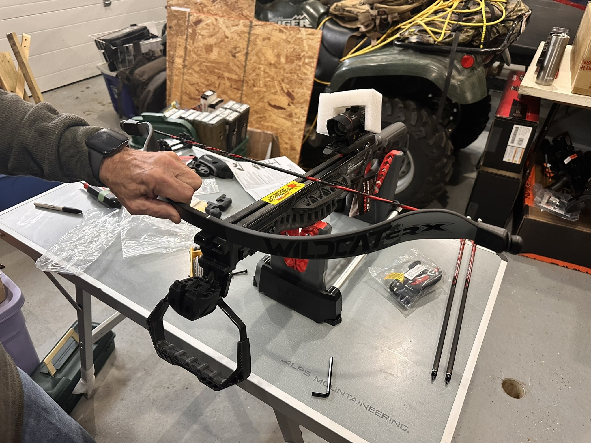 Crossbow mounted on a workbench with a small action camera attached to foam padding on the stock area, in a workshop setting