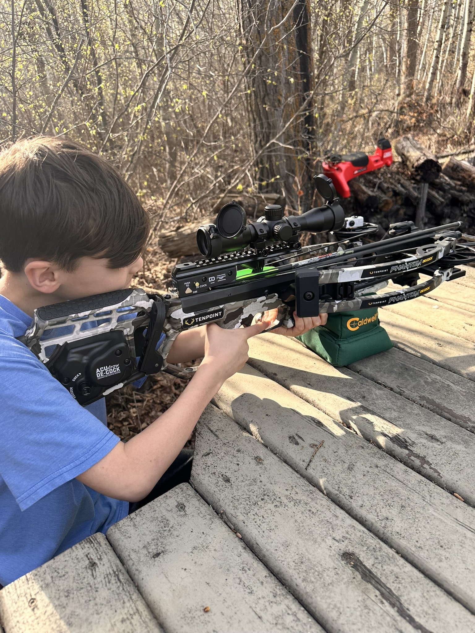 Boy in a blue shirt aims a camouflaged crossbow with a scope from a wooden deck in a wooded area