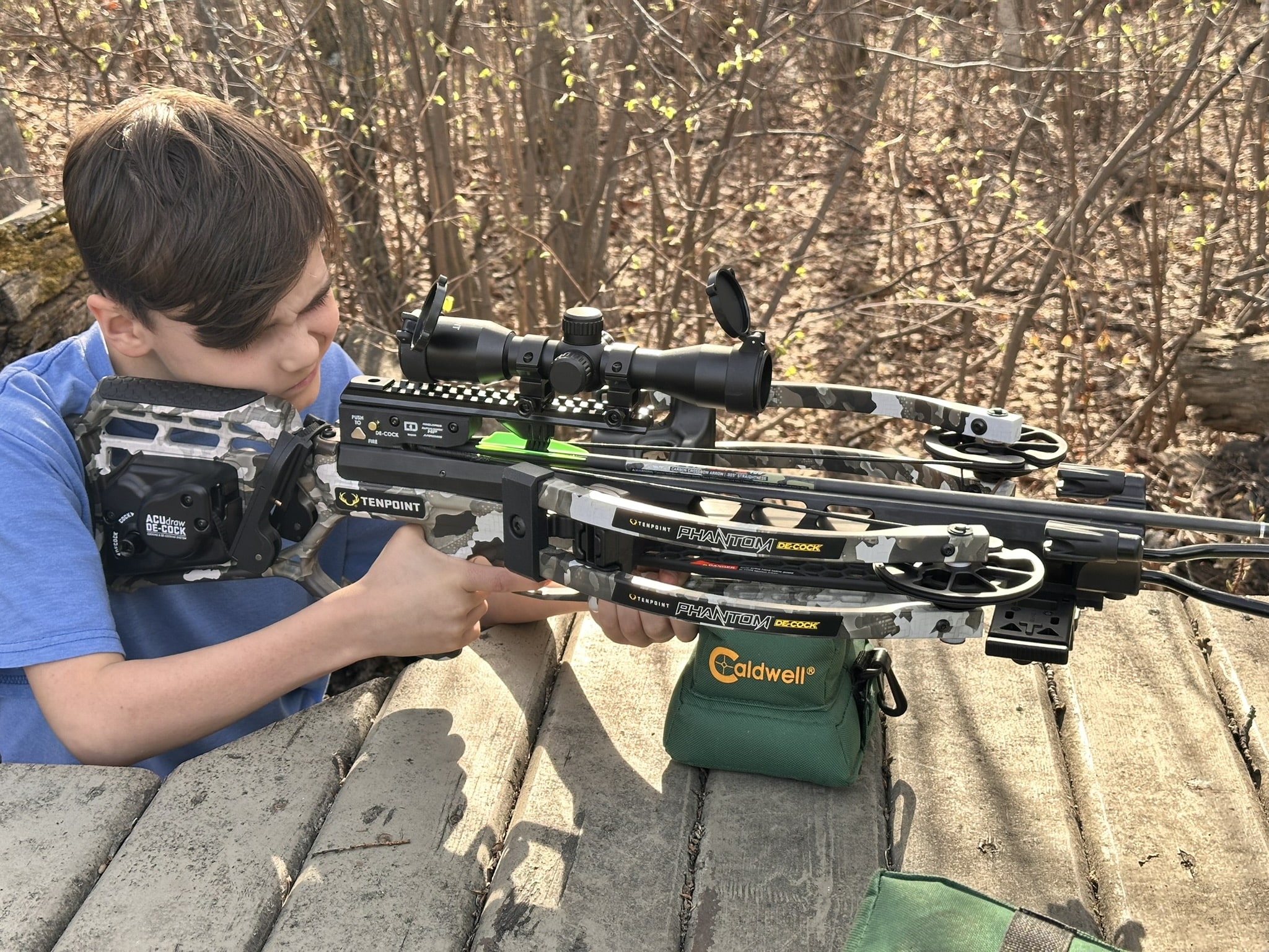 Boy aiming a camouflage crossbow with scope in a wooded outdoor setting.