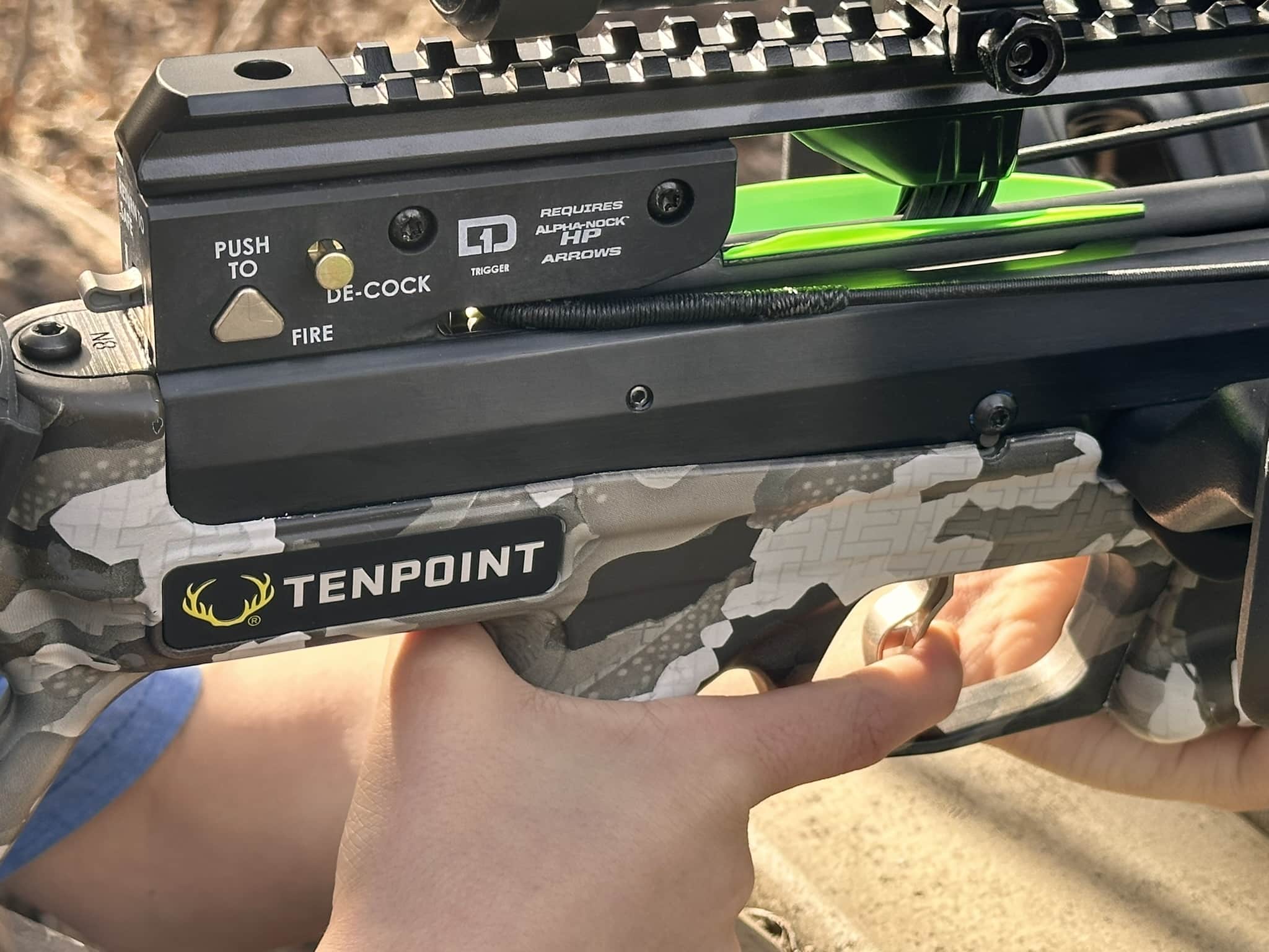 Close-up of a TenPoint camouflage crossbow showing the trigger area and safety labels, with green bolts seated in the limb track.