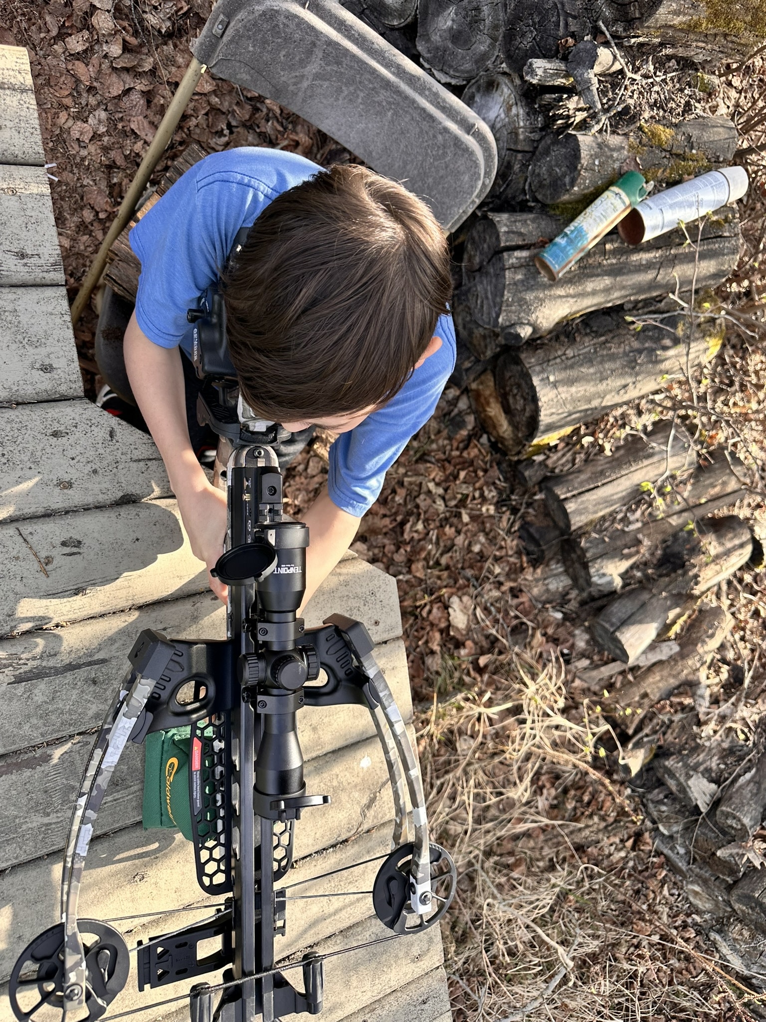 Person in a blue shirt crouches on a wooden deck, assembling a modern crossbow with scope amid fallen leaves and stacked firewood nearby