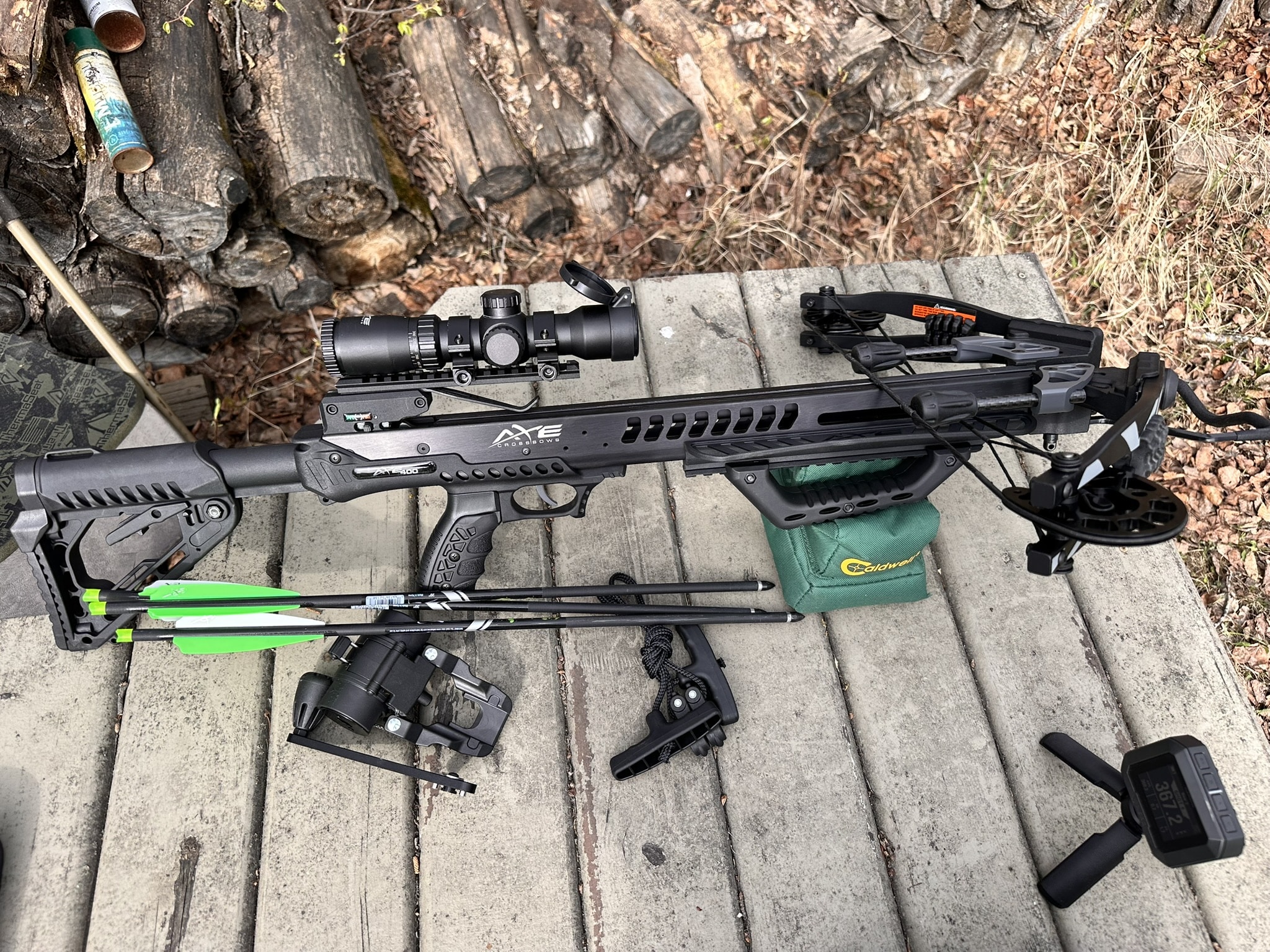 Crossbow with scope and bolts laid out on a wooden surface, along with other archery gear and a green carrying bag nearby