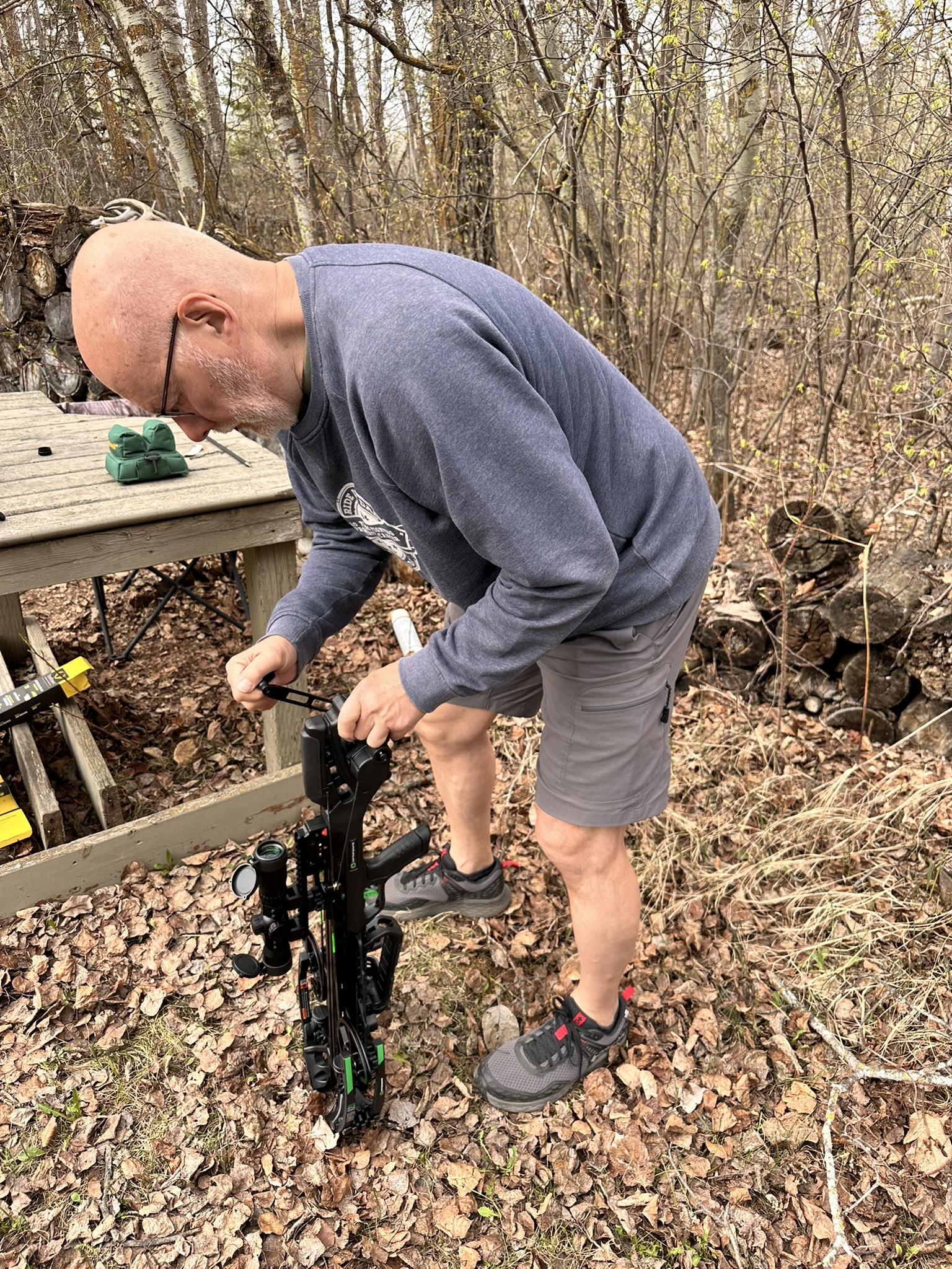 Bald man in a gray sweatshirt adjusts a crossbow outdoors in a leaf-strewn clearing.