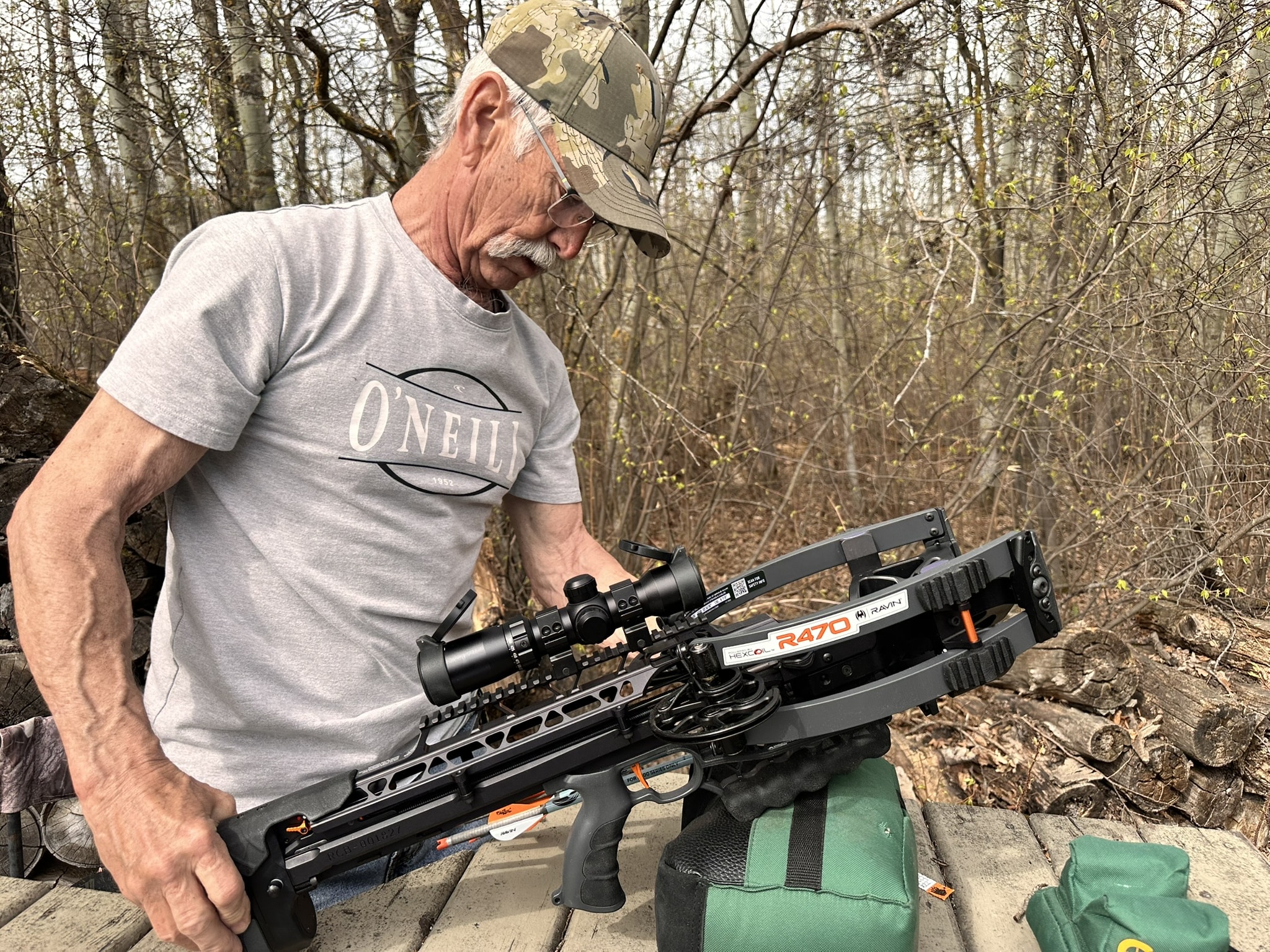 Older man in a camo cap and gray O'Neill shirt handles a large crossbow outdoors among leafless trees, looking down at the weapon.