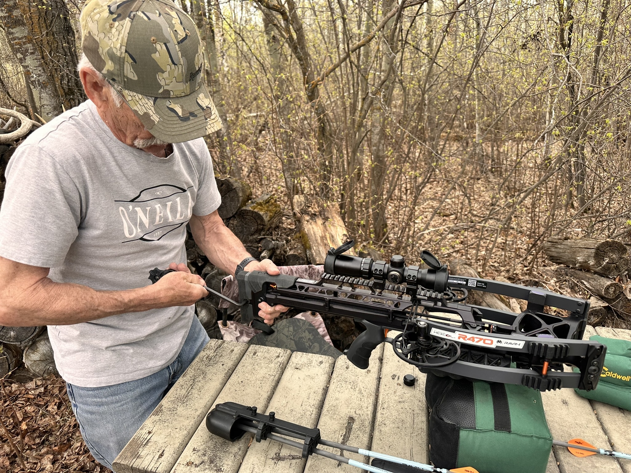 Man wearing a camouflage cap and gray T-shirt, handling a scoped crossbow outdoors in a wooded area.