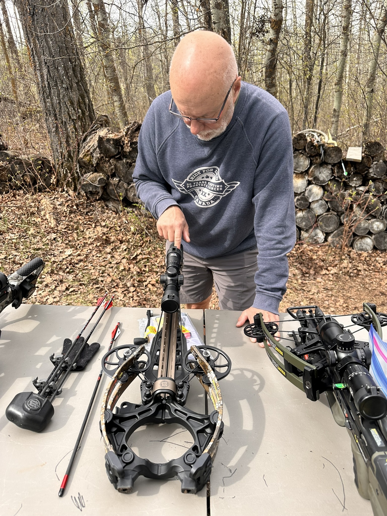 Man adjusting a crossbow on a table outdoors, surrounded by archery gear and arrows.