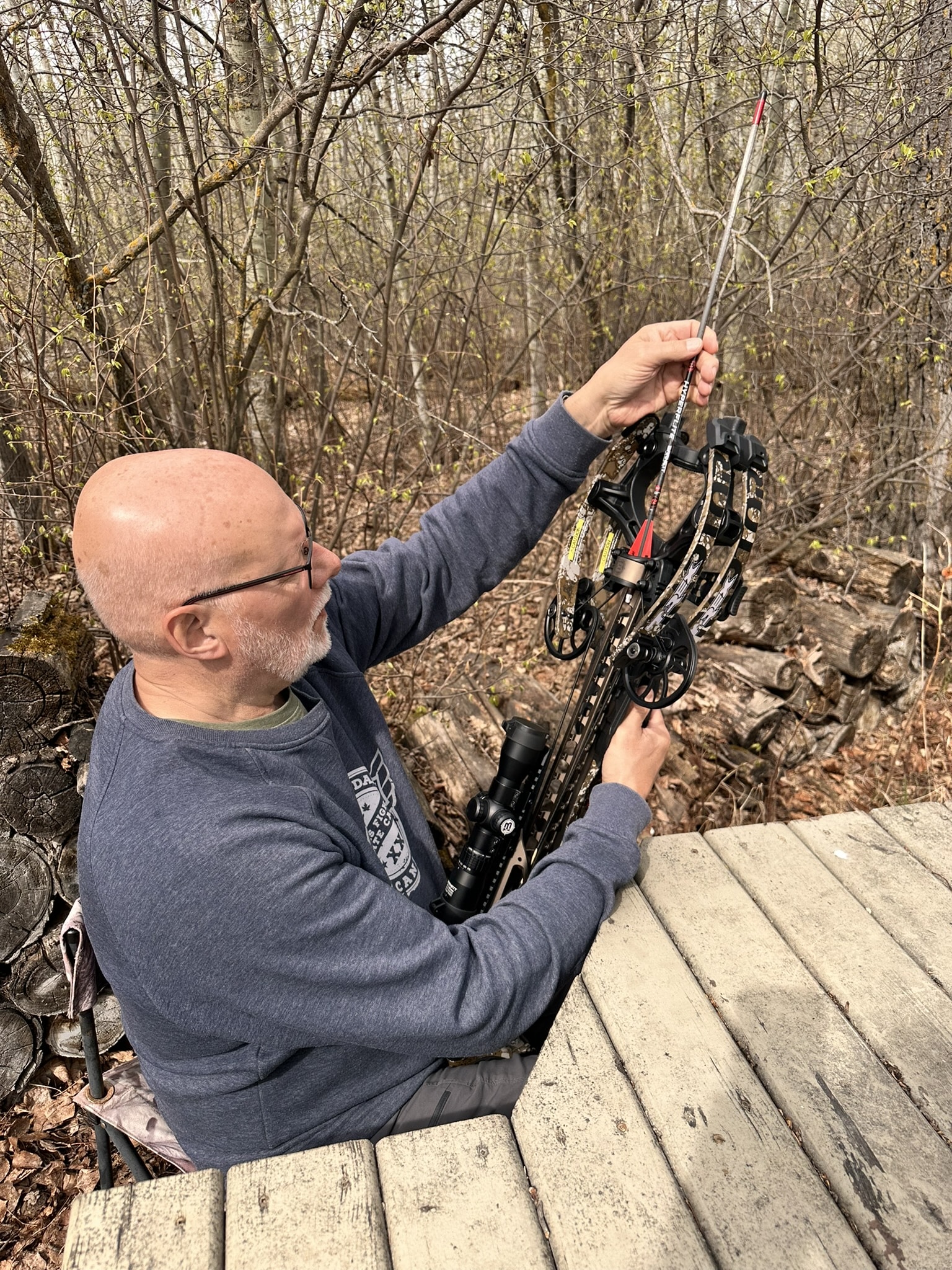 Man with a crossbow and arrows standing on a wooden deck in a forest, preparing to shoot. The archer wears a gray sweatshirt and glasses.