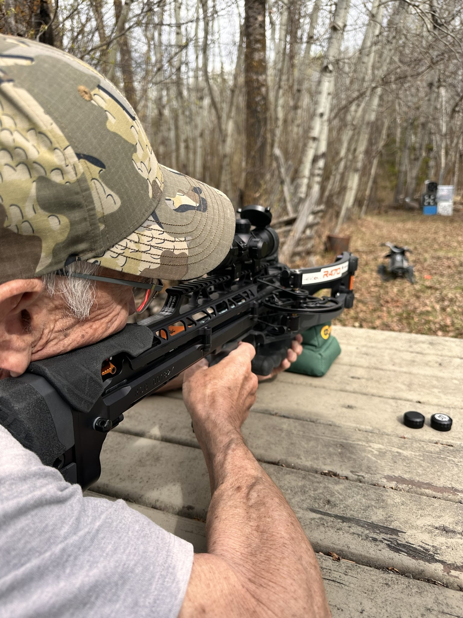 Man in a camouflage cap aims a scoped crossbow from a wooden shooting bench at an outdoor range, with targets visible in the background.