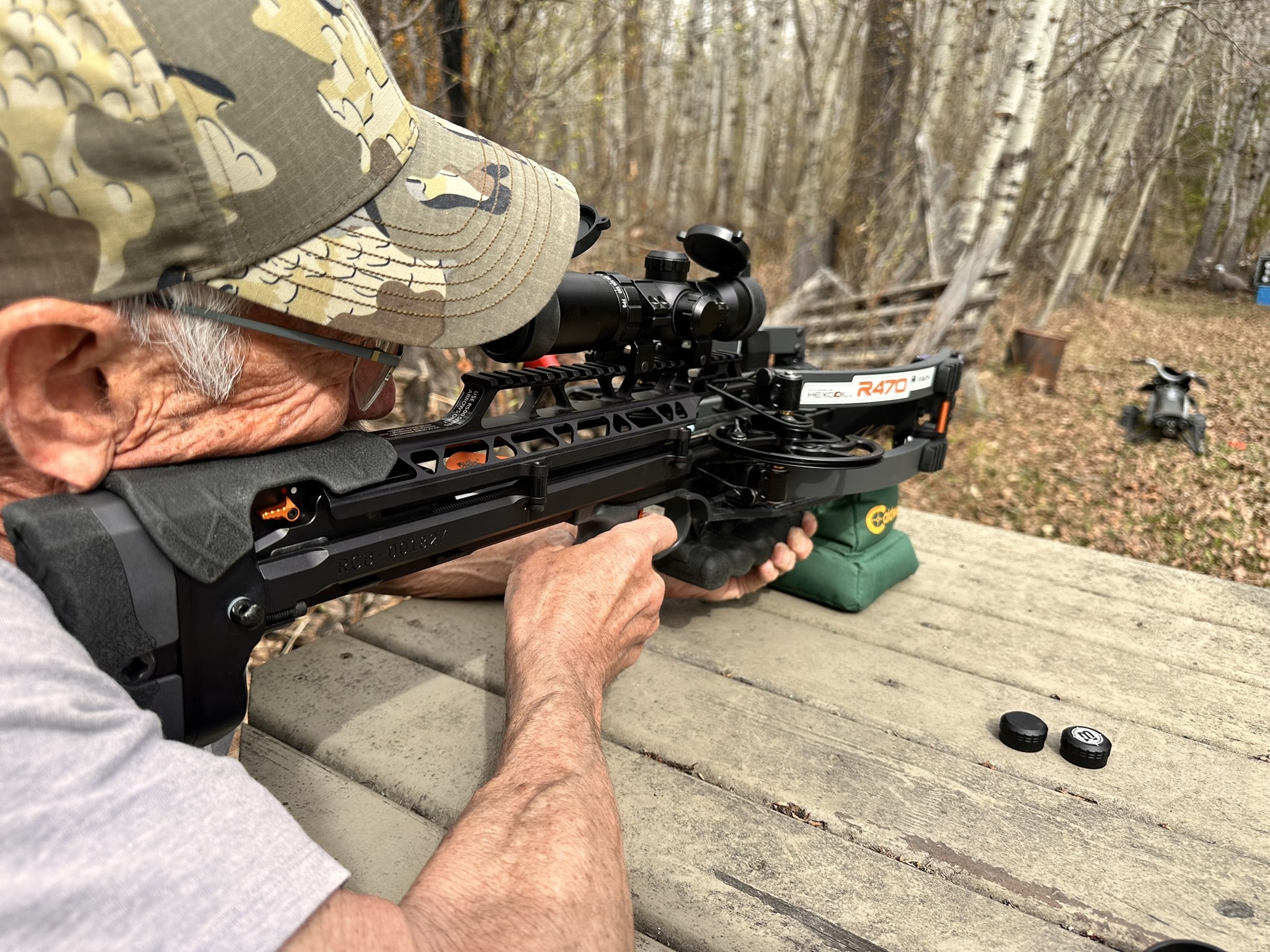 Hunter wearing a camouflage cap aiming a black scope-equipped crossbow in a wooded outdoor setting.