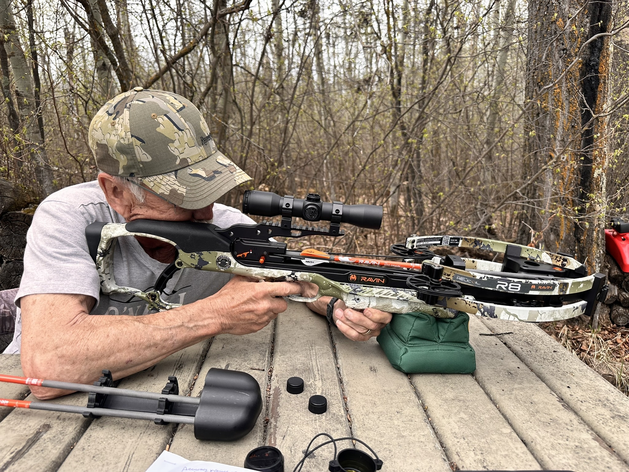 Man in camouflage cap aiming a scoped crossbow outdoors among trees, resting on a wooden bench.