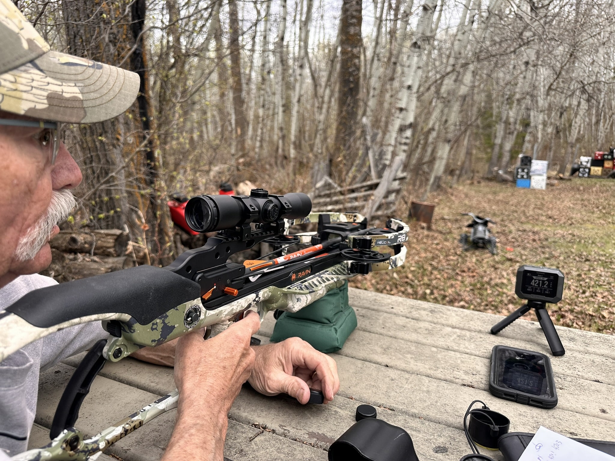 Close-up of a man in a cap aiming a camouflaged scoped crossbow outdoors in a wooded area.