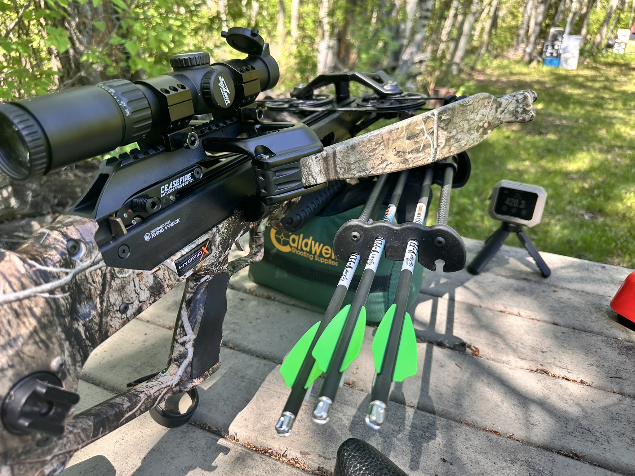 Close-up of a camouflage crossbow with scope and bright green fletched bolts laid on a bench outdoors
