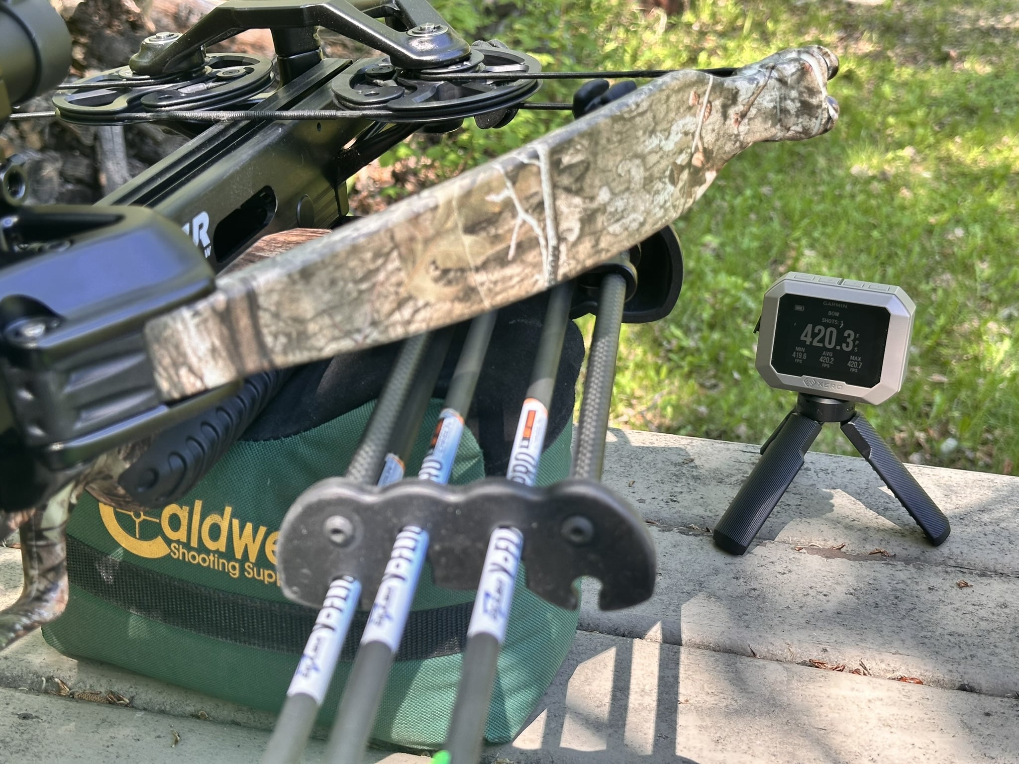 Close-up of a camo hunting crossbow mounted on a green bag with a high-tech bow sight and a Garmin screen nearby outdoors.