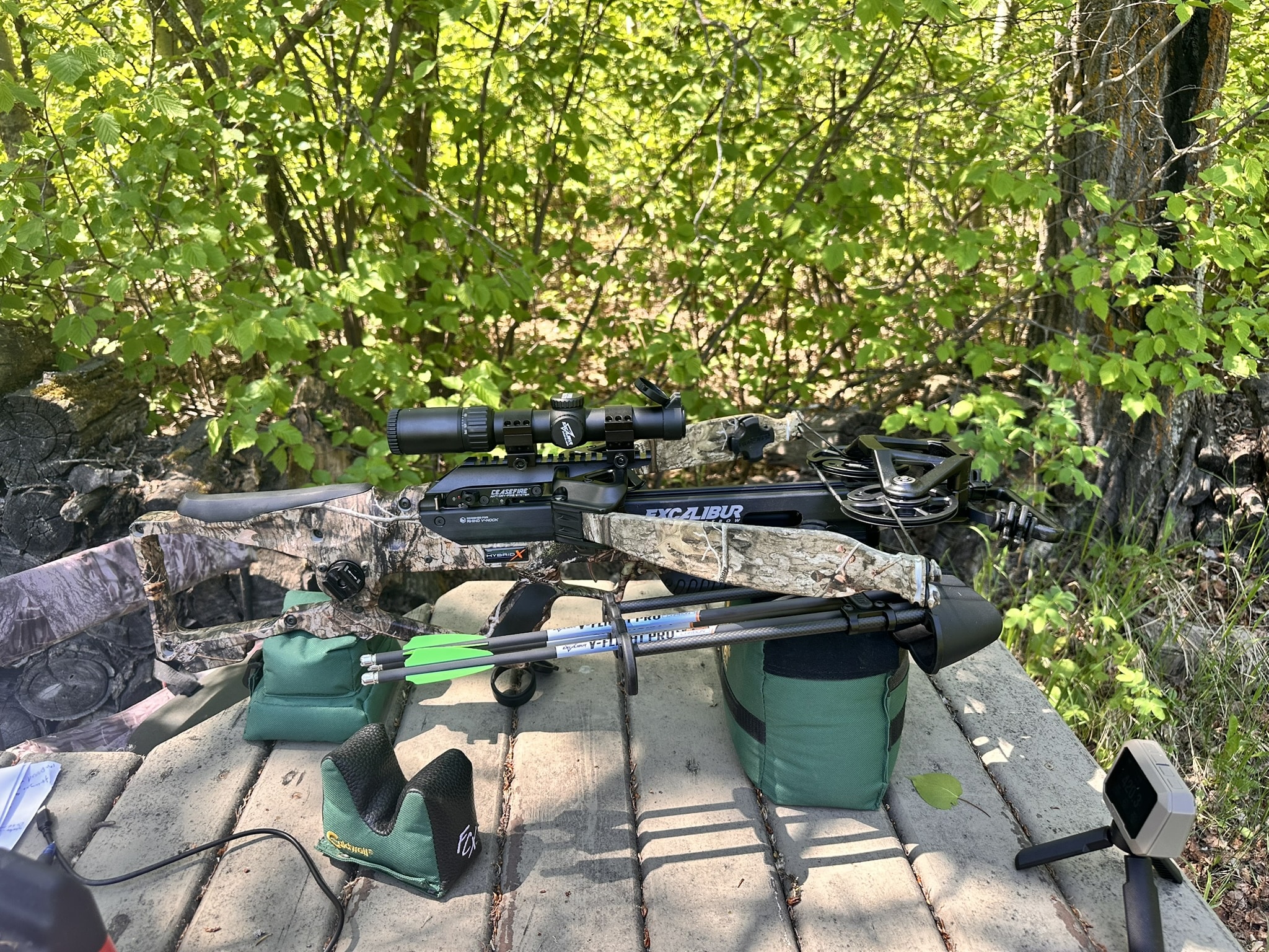 Crossbow with scope resting on a wooden bench outdoors, surrounded by dense green bushes and a quiver of arrows nearby.