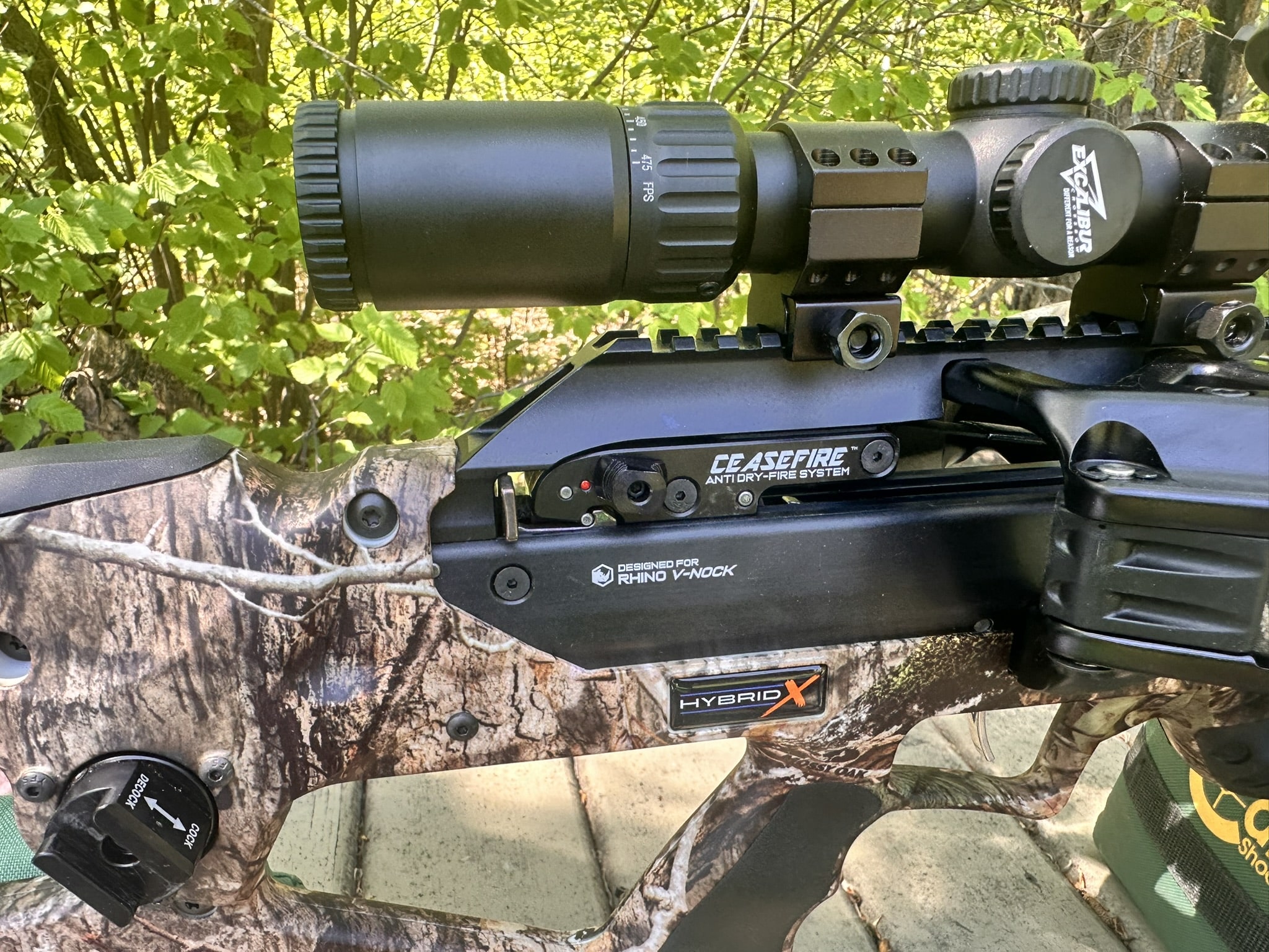 Scope mounted on a camouflage crossbow with CeaseFire anti dry-fire system and Rhino V-Nock branding visible, outdoors among leaves.