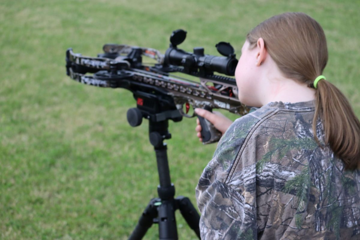 Person in camouflage aiming a modern scoped crossbow on a bipod in a grassy field.
