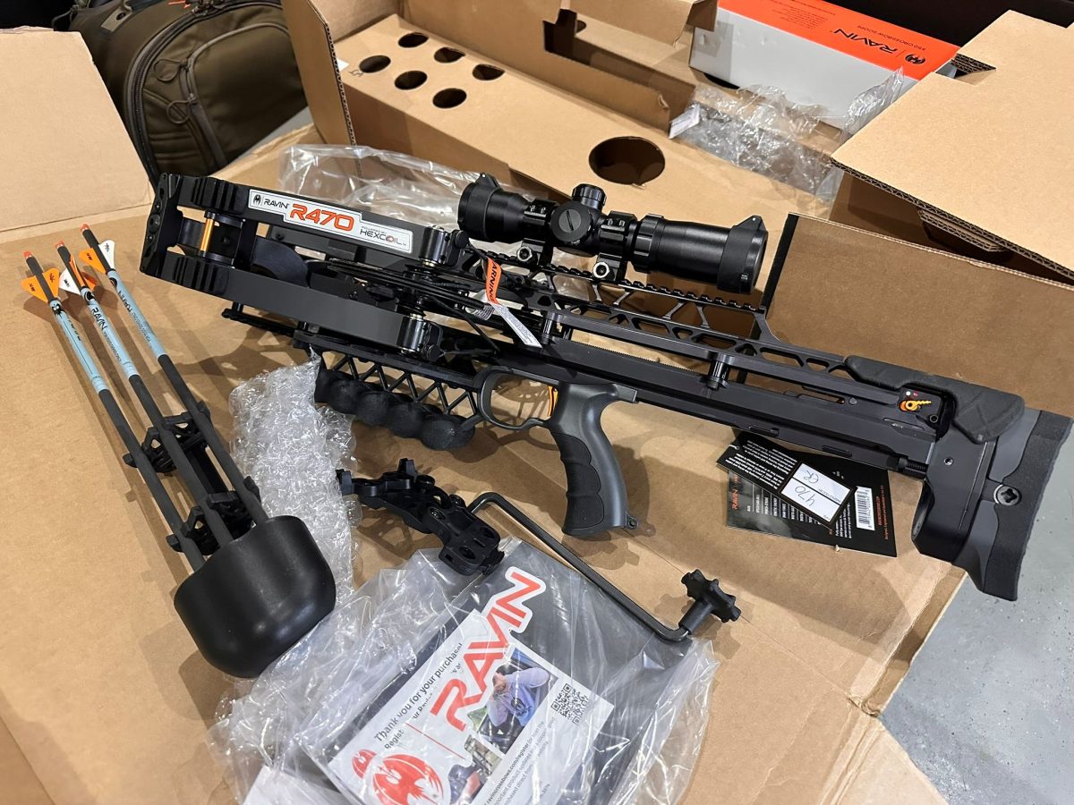 Ravin R470 crossbow with scope and orange-fletched arrows in a cardboard box, surrounded by packing material and accessories.