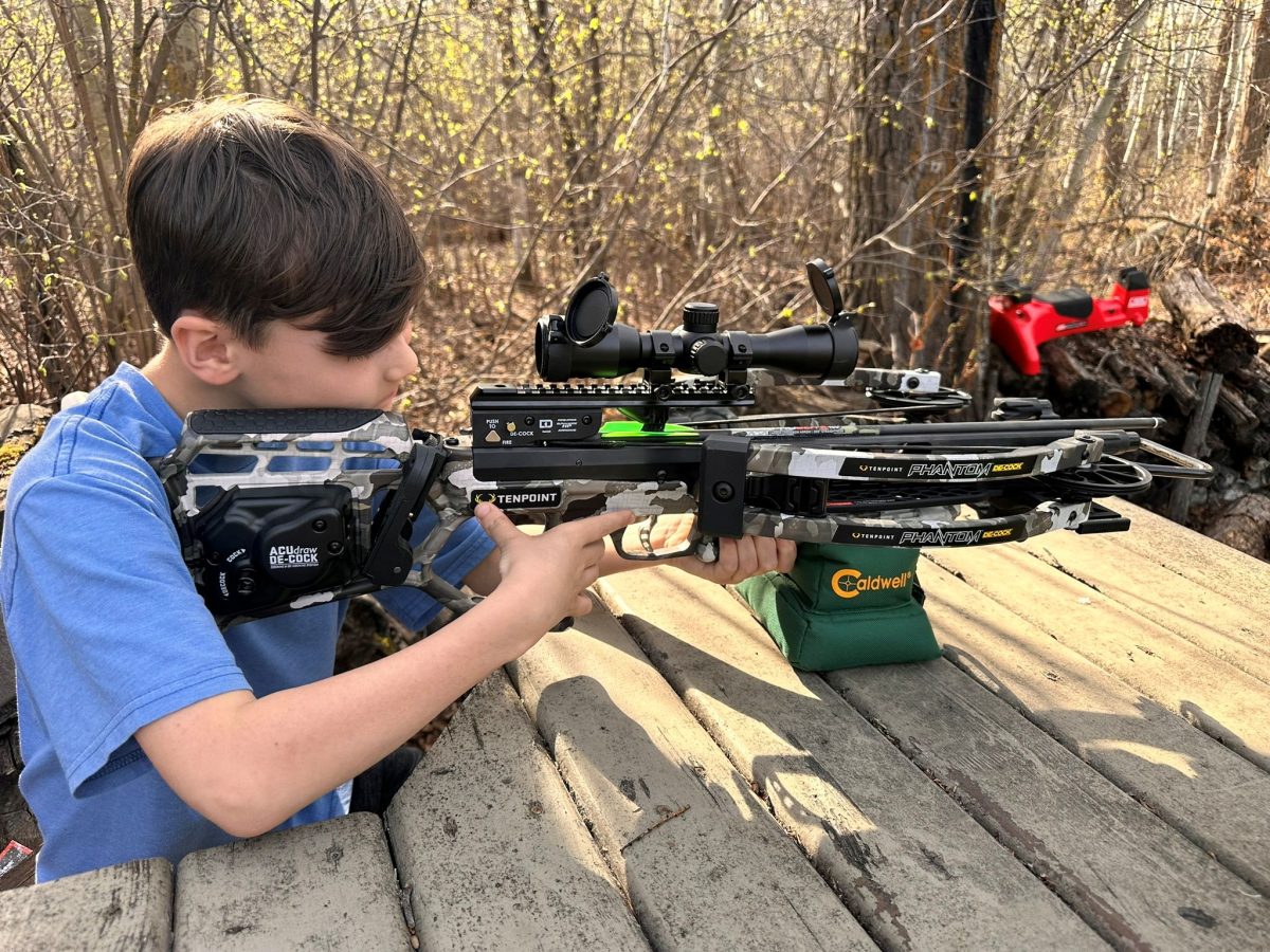 Young boy in a blue shirt aiming a camouflaged crossbow with a scope on a wooden outdoor platform, forest in the background.