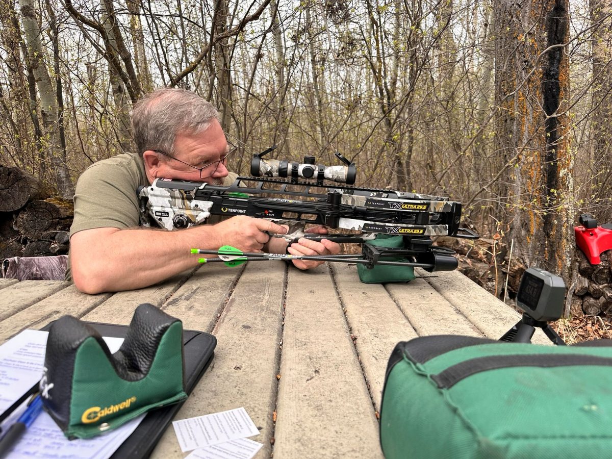 Man lying prone in a forest, aiming a camouflaged crossbow with a scope, surrounded by archery gear and bags.