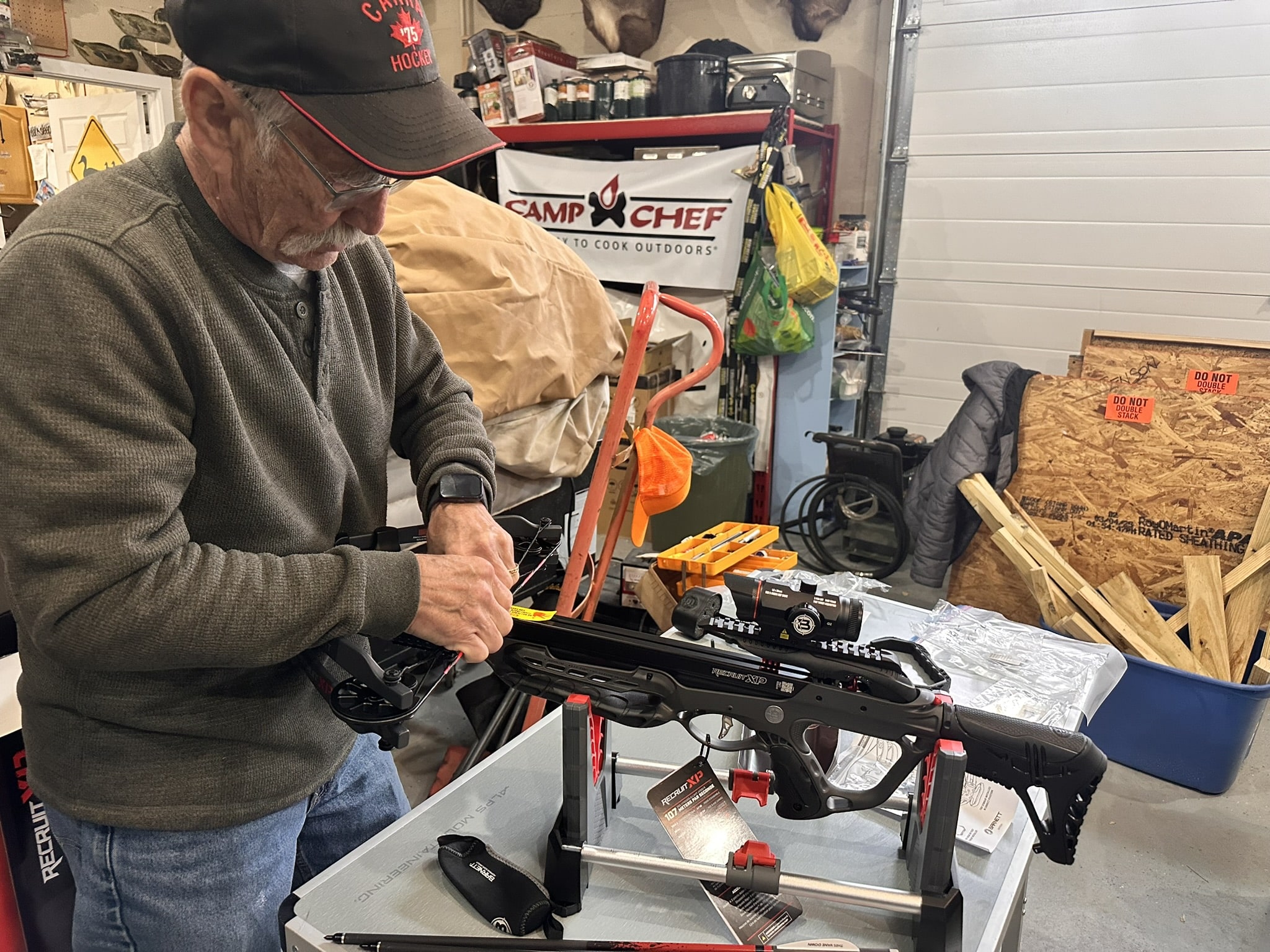 Older man wearing a dark cap and glasses works on a crossbow in a cluttered workshop filled with tools and gear at a workbench.