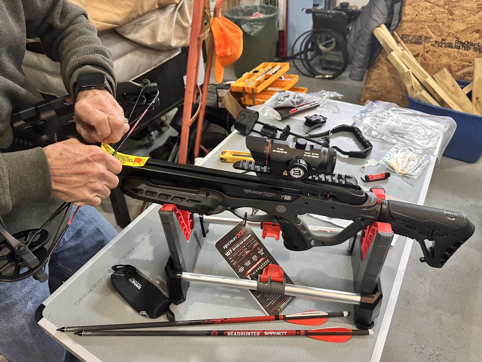 Person assembling a crossbow on a workbench with tools and orange gloves in the background.