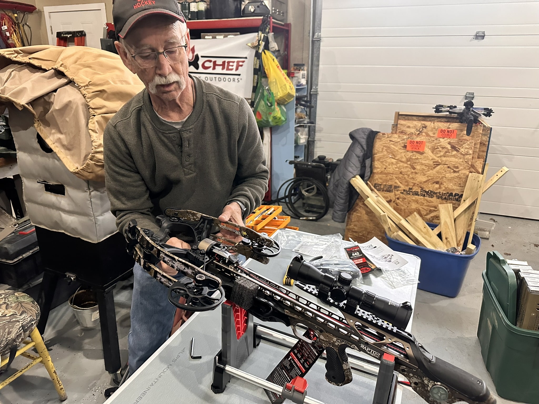 Older man wearing a cap and glasses inspecting a camo-pattern hunting crossbow at a cluttered workshop workbench.