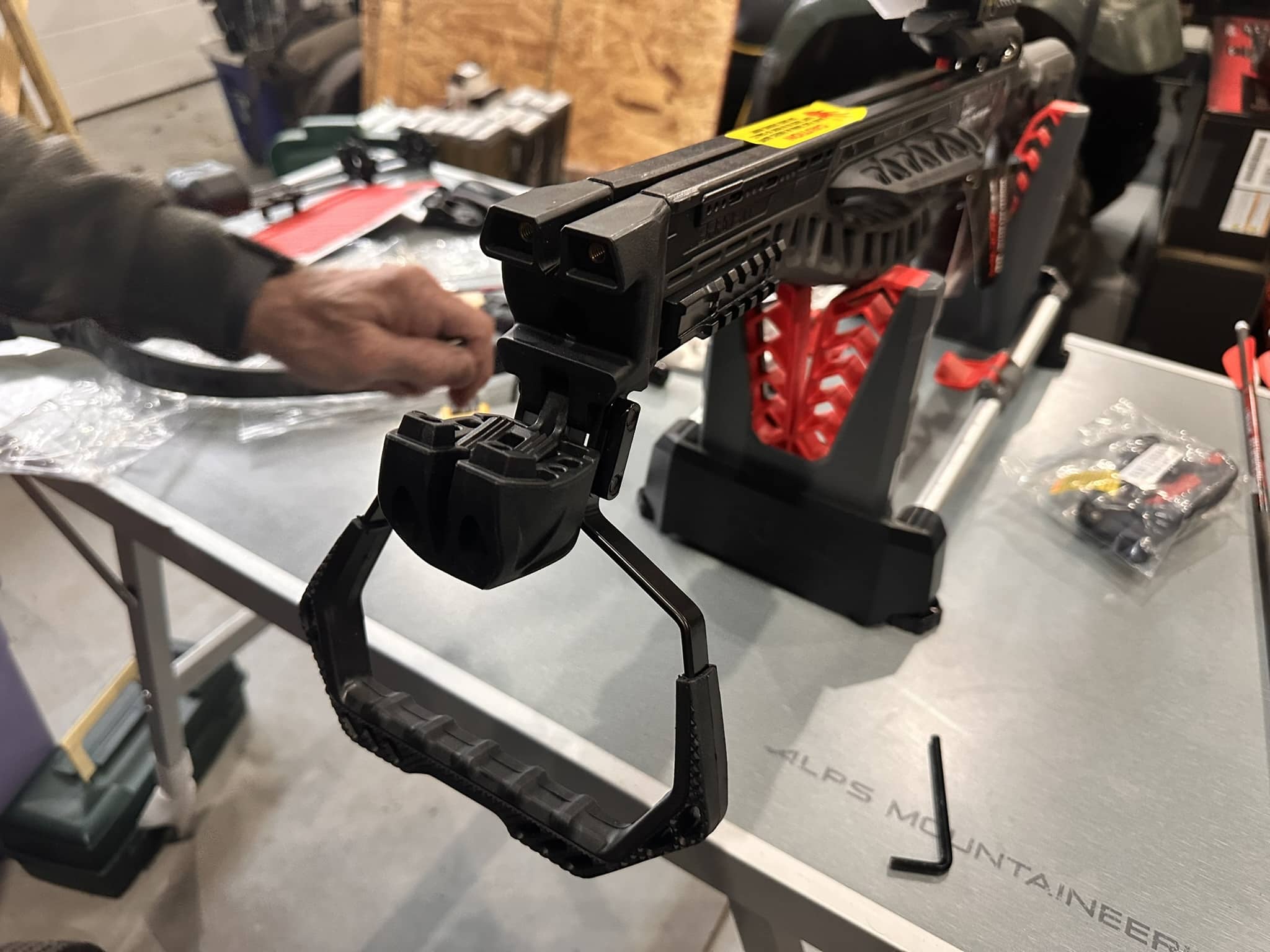Close-up of a black rail-equipped crossbow mounted on a stand with a person’s hand nearby on a workshop table