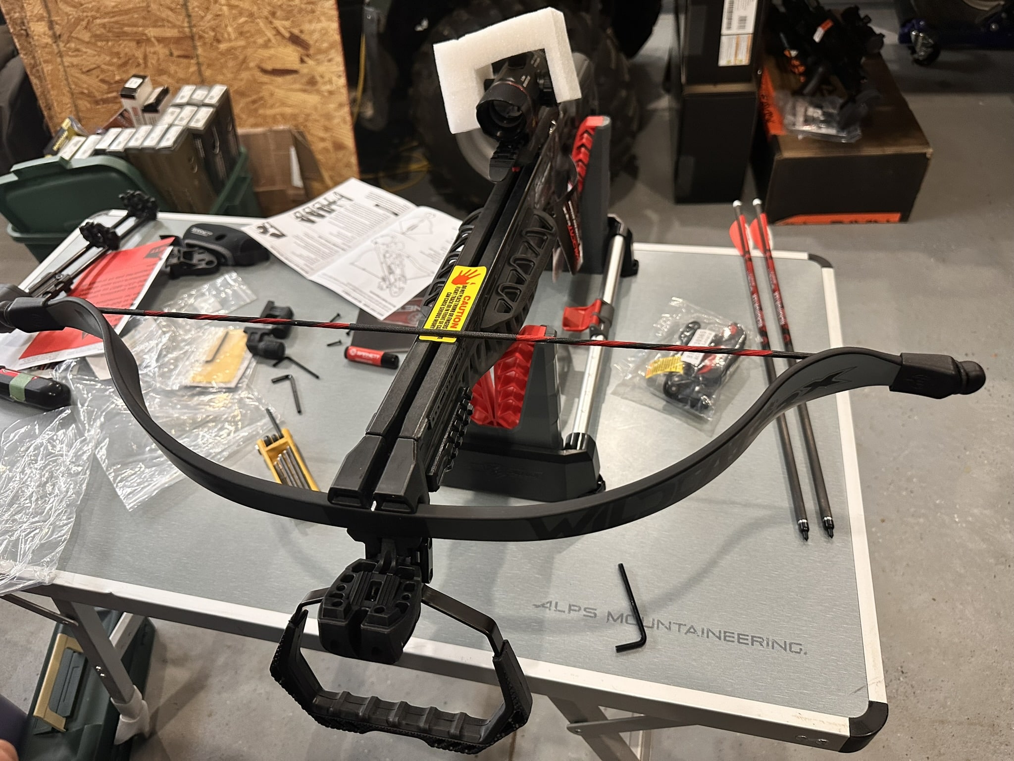 Close-up of a modern recurve crossbow on a workbench with tools, manuals, and packaging in the background.