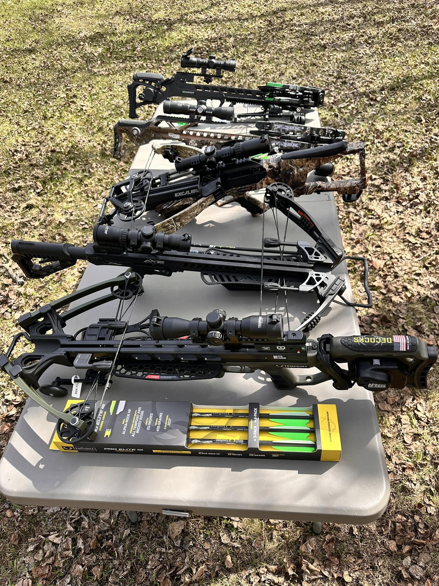 Several modern hunting crossbows with scopes and bolts laid on a gray folding table outdoors.