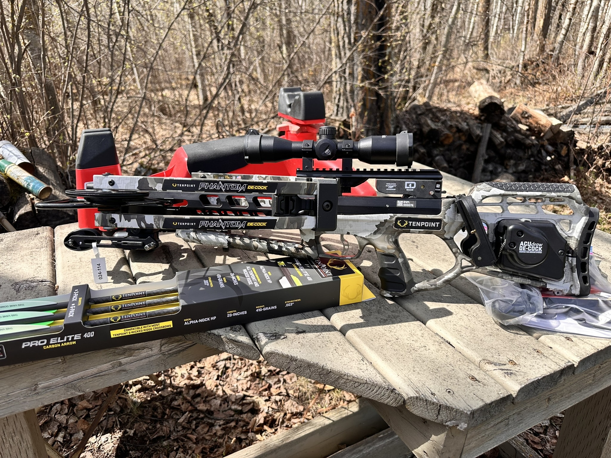 TenPoint Phantom De-Cock crossbow with scope mounted, sitting on a wooden table outdoors among boxes of bolts and arrows.