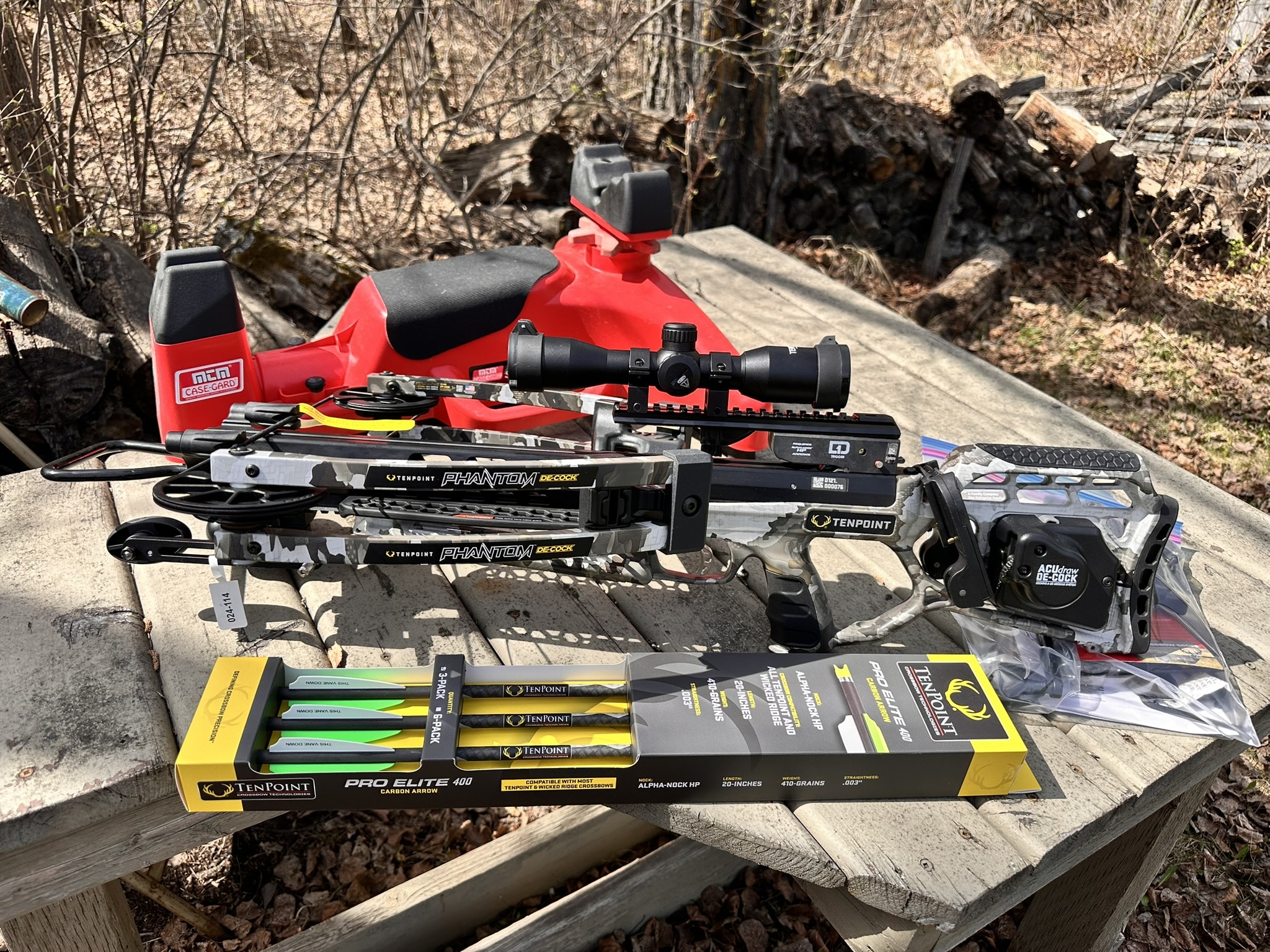 Crossbow with a scope and accessories laid on a wooden bench outdoors, with yellow/green TenPoint arrows nearby.