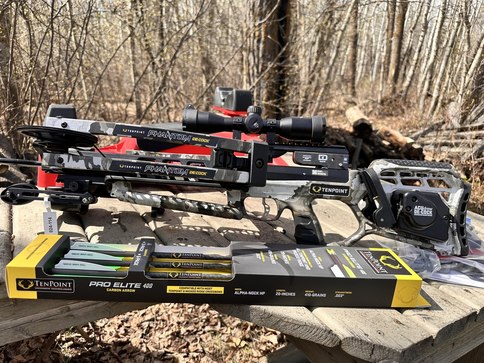 Crossbow setup on a wooden bench with camouflage limbs and a scope mounted, outdoors in a wooded area.