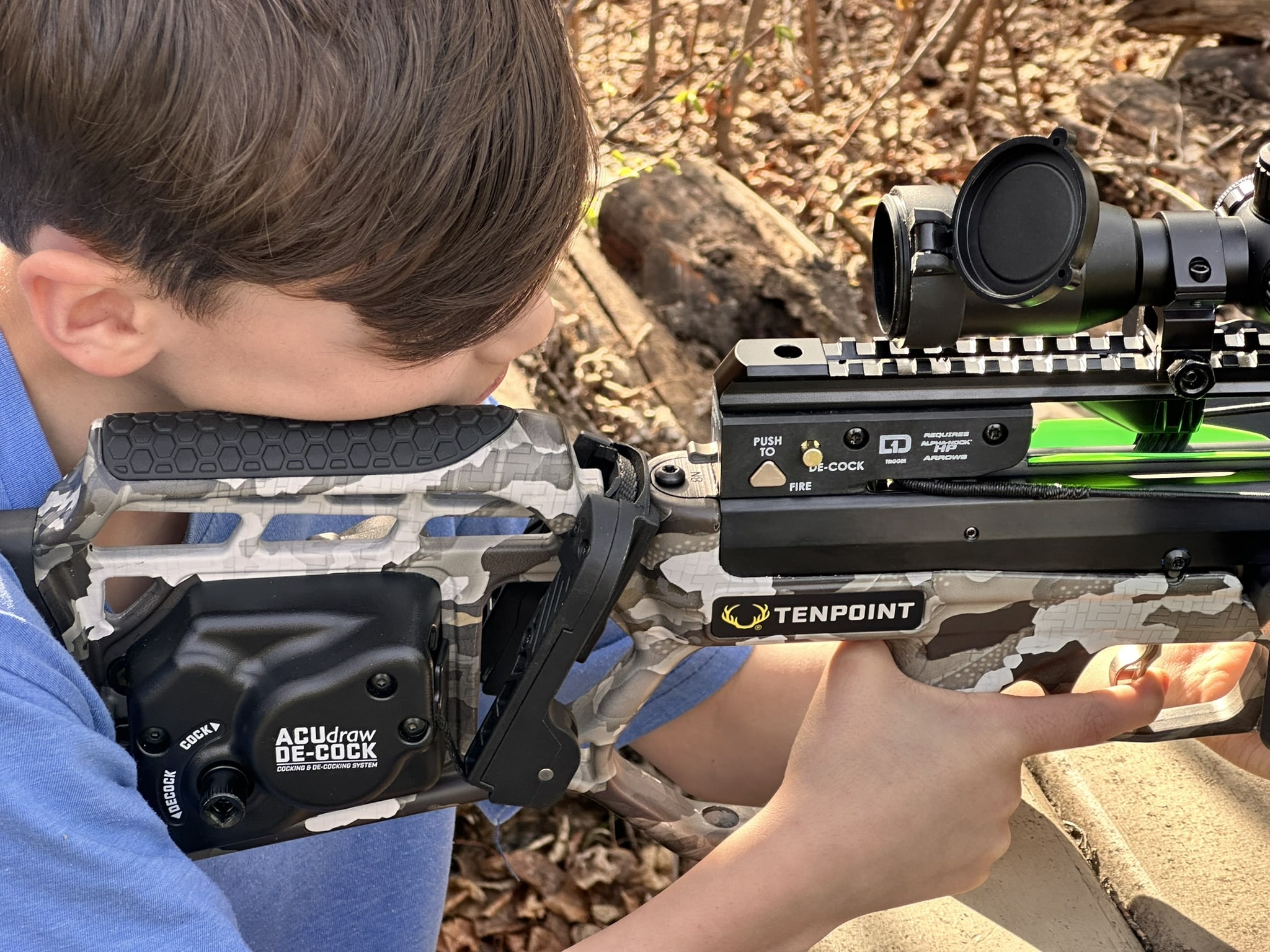 Boy in camo clothing aiming a TenPoint crossbow with scope outdoors in a wooded area.