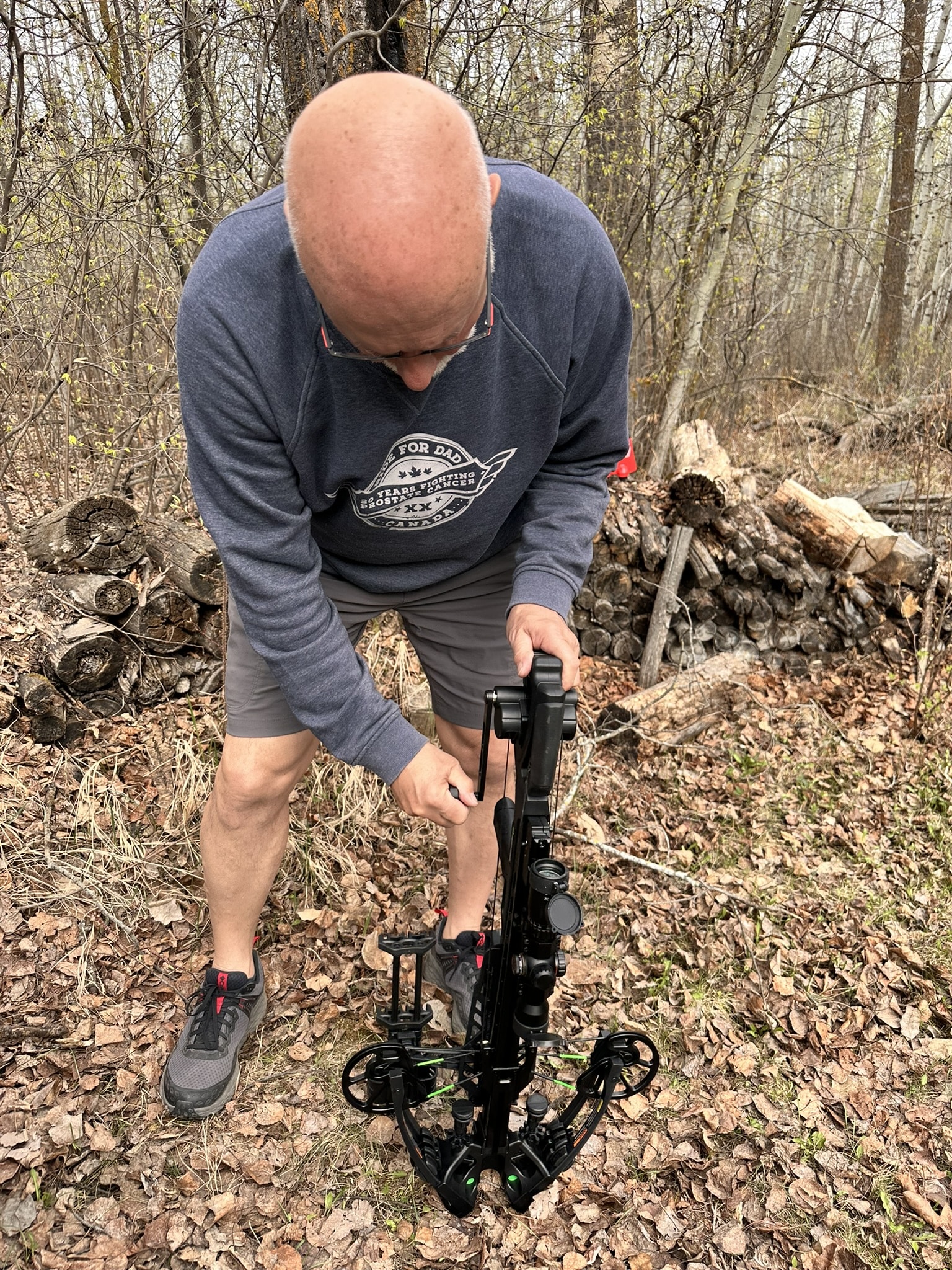 Man bending over to adjust a crossbow bow in a leaf-covered forest clearing, wearing a blue sweatshirt and shorts.