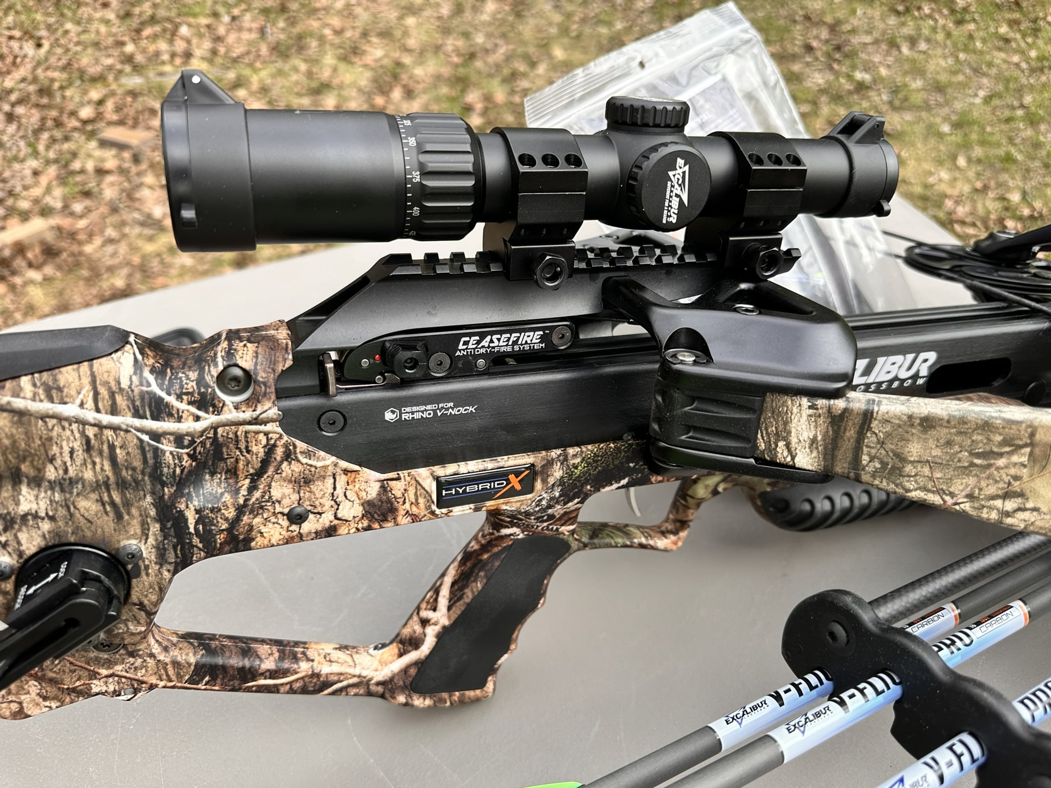 Camouflage crossbow with a large scope mounted on a rail system, resting on a wooden surface outdoors